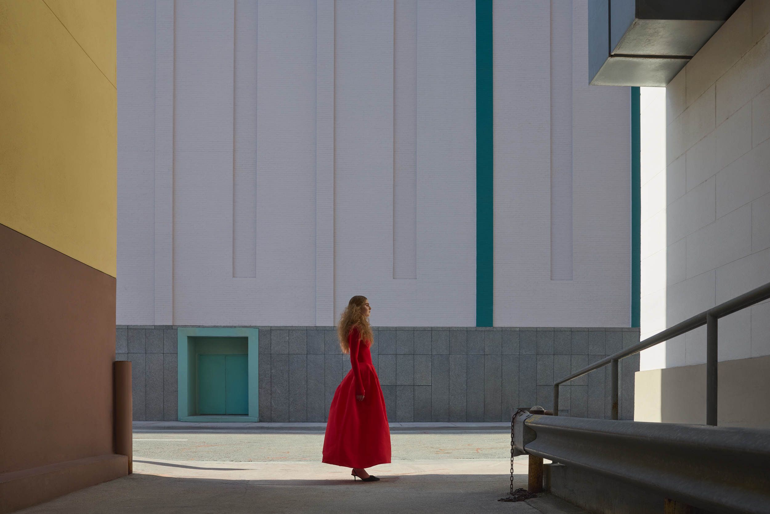Female model in a red dress standing within geometric modern architecture from the Farewell fashion editorial by Melina Deya